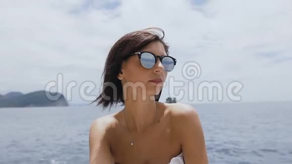 美丽的女孩坐在岩石上戴着太阳镜穿着白色泳衣背景是深蓝色的海水亚得里亚海视频的预览图