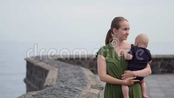 一位年轻的妈妈带着一个婴儿在她的手上射击看着海洋和海浪的惊人景色显示孩子视频的预览图