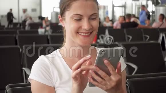 年轻女子坐在机场候机室里带着手机女性旅行者浏览智能手机视频的预览图