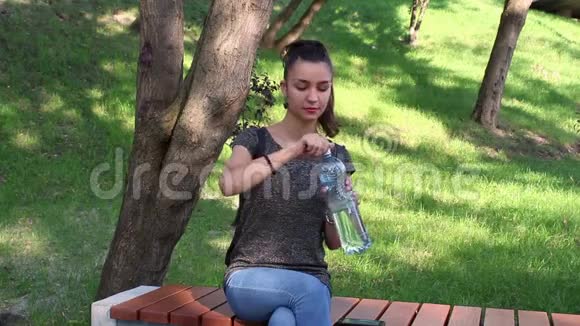 年轻漂亮的女孩坐在公园的长凳上喝水视频的预览图