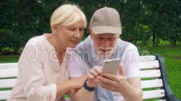 老两口在公园里坐在长凳上聊天放松在智能手机上浏览视频的预览图