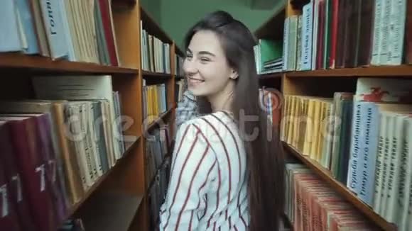 美丽的女孩留着长长的黑发走到图书馆的书架之间手里拿着书慢动作广泛视频的预览图