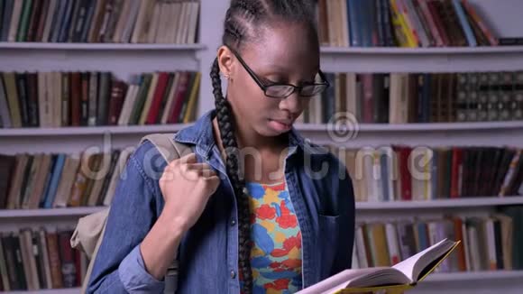 年轻美丽的非洲裔美国妇女戴着眼镜在图书馆看书看着相机开心地笑着视频的预览图