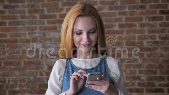 年轻可爱的金发女孩正在智能手机上输入信息看着相机微笑砖背景交流视频的预览图