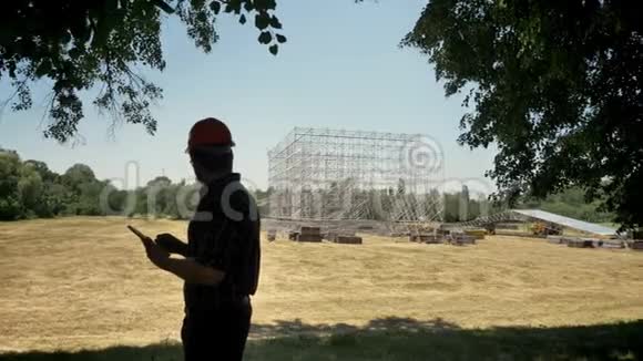年轻建筑师戴着头盔使用平板电脑看着新的金属建筑规划新项目视频的预览图