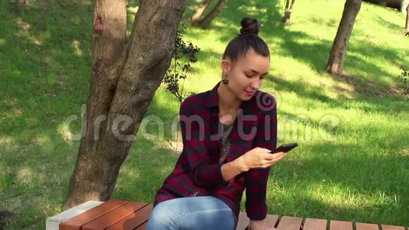 年轻漂亮的女孩穿着格子衬衫微笑着浏览智能手机上的社交网络磁带视频的预览图