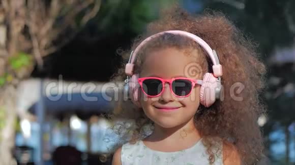 一个可爱的孩子的肖像一个穿着粉色眼镜和粉色耳机的白色裙子的美丽的小女孩视频的预览图