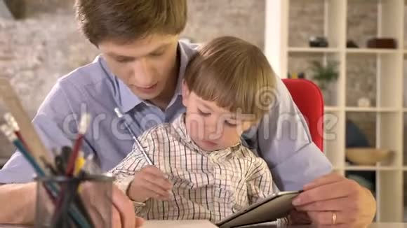 年轻的父亲抱着他的小儿子坐在桌子旁写着纸现代的办公背景视频的预览图