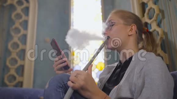 戴眼镜的年轻女子坐在一家咖啡馆里里面有一个亚洲人她抽着烟在智能手机里上网视频的预览图