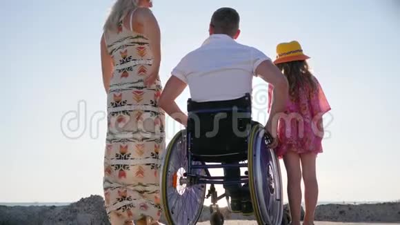 幸福的一家人举起双手看波浪后景丈夫坐轮椅拥抱妻子和小女孩残疾人拥抱视频的预览图
