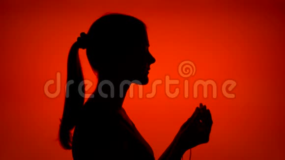 年轻女性的剪影戴上耳机在红色背景下用智能手机听音乐视频的预览图