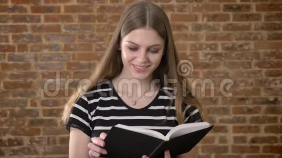 年轻漂亮的金发女郎正在看书看着镜头微笑着带着砖头背景视频的预览图