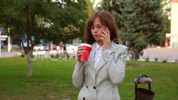 漂亮的女商人戴着轻便的眼镜在公园里喝咖啡用智能手机聊天视频的预览图