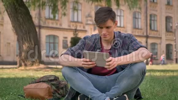 年轻帅哥坐在公园的草地上在平板电脑上看网页在镜头前看放松概念视频的预览图