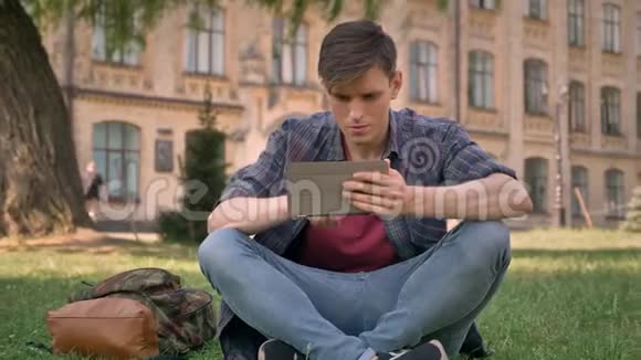 年轻人坐在公园的草地上看着平板电脑上的网页看着相机放松自己的观念视频的预览图