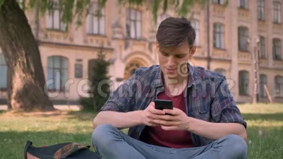 年轻的帅哥坐在公园的草地上敲着智能手机放松概念交流概念继续前进视频的预览图