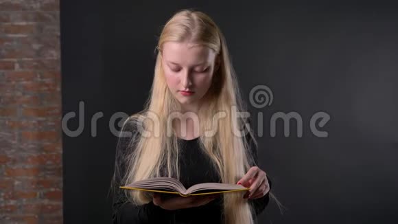 年轻漂亮的金发女孩涂着粉红色口红正在看书看着相机微笑着灰色的背景视频的预览图