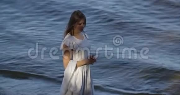 美丽的黑发白种人女人穿着浅色的白色连衣裙站在海浪的背景上思考吸引力视频的预览图