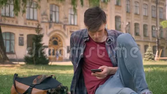 年轻的帅哥坐在公园的草地上看着智能手机上的互联网页面放松概念交流视频的预览图