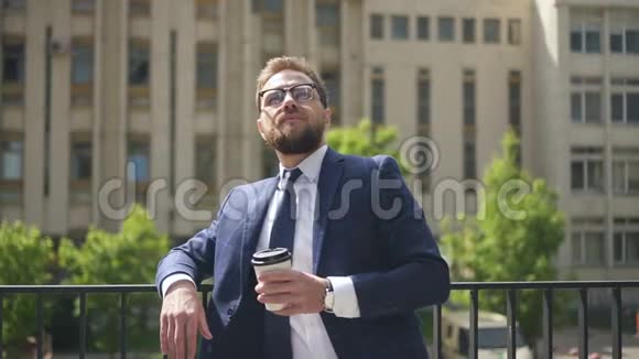 年轻的胡子商人拿着咖啡杯看着站在城市街道上的手表视频的预览图