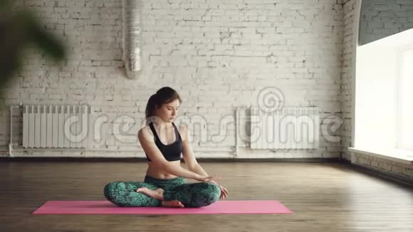年轻的女士穿着时髦的运动服正在做身体扭曲坐在莲花的位置在瑜伽工作室里的垫子上放松一下视频的预览图