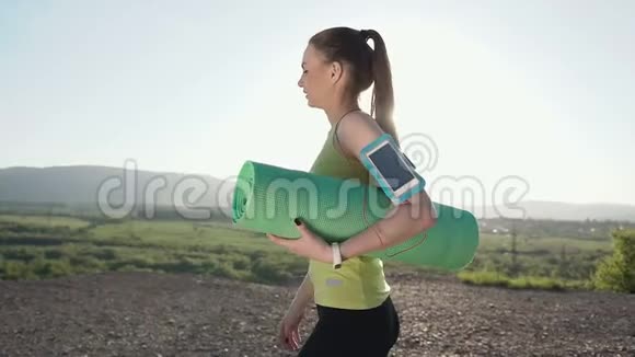 年轻的运动女子步行在日落时做晚间练习女孩拿着绿色瑜伽垫女运动员穿过视频的预览图