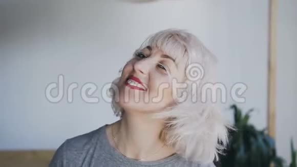 一个疯狂的金发女郎在白色的墙壁背景下摇着头摇着头发的特写肖像视频的预览图
