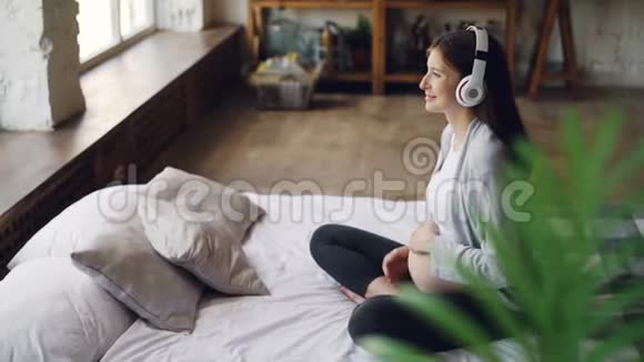 微笑的怀孕女孩正在听耳机音乐抚摸她的腹部放松地坐在双人床上视频的预览图