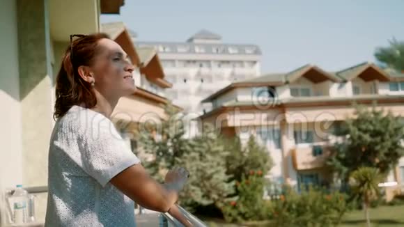 一个穿着白色连衣裙的女人在阳台上享受温暖的阳光视频的预览图