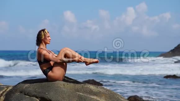 一个穿比基尼的年轻女孩坐在石头上交替地举起腹部肌肉做运动视频的预览图