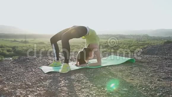 穿着紧身衣的年轻女子在日出或日落时在山顶练习瑜伽健身体育视频的预览图