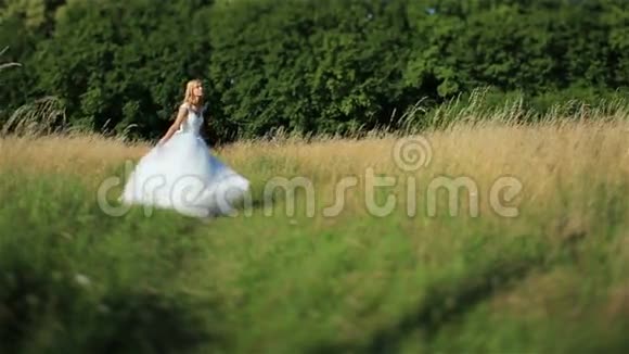 美丽浪漫时尚的金发新娘穿着白色连衣裙在阳光下在绿色的田野里跳舞视频的预览图