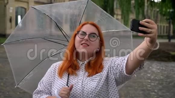 戴着眼镜的胖姜女孩正在公园里的智能手机上在雨天自拍在公园里闲逛拿着雨伞视频的预览图