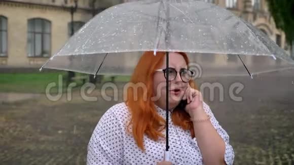 戴眼镜的肥姜女孩站在公园的雨下打电话撑着伞交流理念视频的预览图