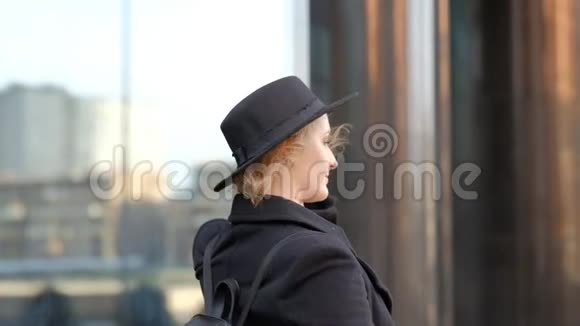 美丽的金发女郎戴着黑色帽子微笑着摆姿势对着镜头向上看着建筑近距离观看慢动作视频的预览图