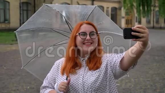 戴着眼镜的肥姜女孩正在公园里的智能手机上在雨天自拍到处闲逛拿着雨伞视频的预览图