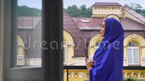 年轻迷人的穆斯林妇女戴着头巾站在阳台上欣赏美丽的景色视频的预览图