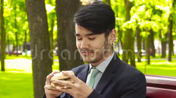 亚洲年轻商人正坐在公园的长椅上嗅着汉堡视频的预览图
