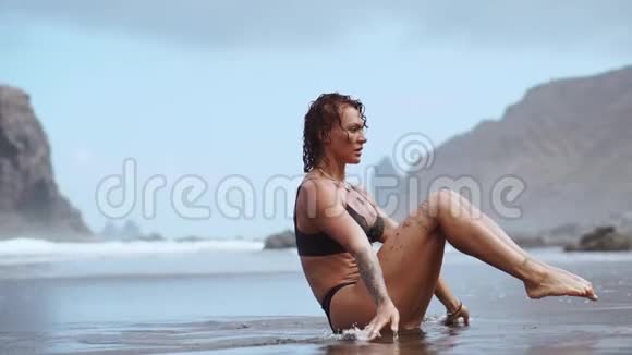 运动女性在沙滩上训练腹部肌肉在岩石和海浪的背景下用黑色沙子训练视频的预览图