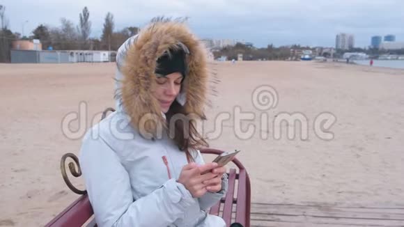 穿着白色羽绒服的女人坐在海滩上的长凳上看着手机视频的预览图