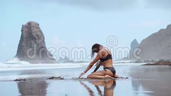 美丽的女人穿着泳装在海滩上跳舞沙滩上有黑色的沙子靠近大海视频的预览图