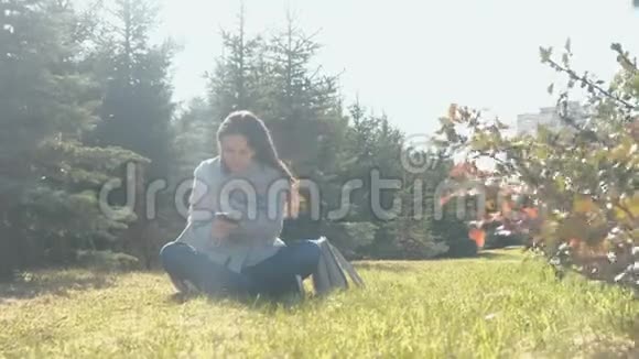 年轻美丽的黑发女子坐在草地上的春风公园并键入一个信息在手机视频的预览图