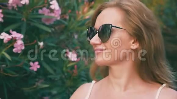 美丽时尚的金发女人戴着太阳镜带着开花的灌木在公园里散步夹竹桃花反映在视频的预览图