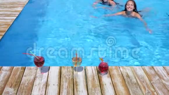 三位穿比基尼的年轻女士喝着五颜六色的鸡尾酒在游泳池的蓝色水中放松视频的预览图