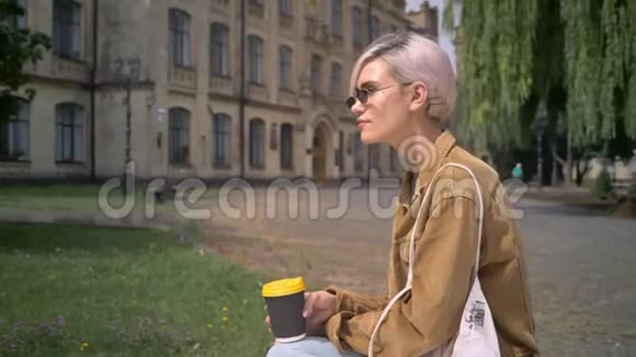 在大学附近坐着喝咖啡戴着金色短发眼镜的时尚女性视频的预览图