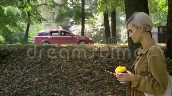 年轻的美女短发手里拿着咖啡打着电话走在公园里视频的预览图