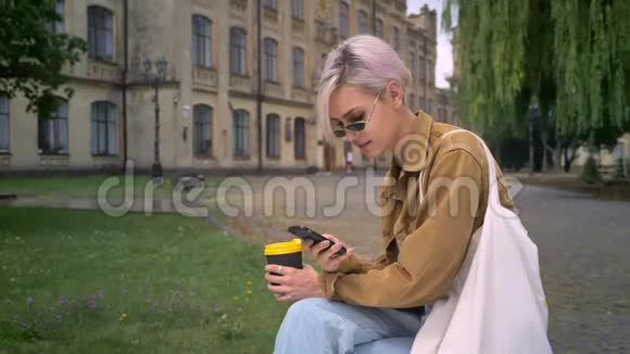 年轻的金发女郎戴着太阳镜短发端着咖啡打着电话坐在大学附近视频的预览图