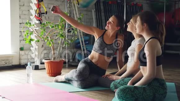 瑜伽的学生们正在用智能手机自拍同时坐在瑜伽工作室里明亮的垫子上女人摆姿势视频的预览图
