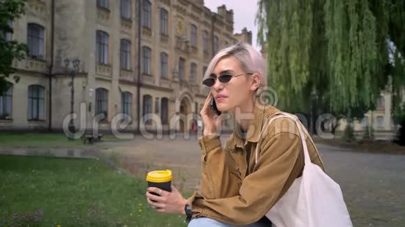 穿着墨镜的时髦漂亮女人拿着咖啡和电话坐在大学附近的公园里视频的预览图