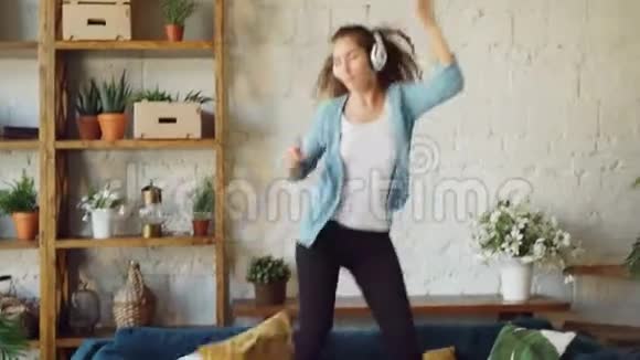 漂亮的黑发女郎在家里玩得很开心通过耳机听音乐在沙发上唱歌跳舞视频的预览图